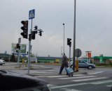 Na fotografii widać przejście dla pieszych z sygnalizacją świetlną. Na czerwonym świetle, przez pasy, przechodzi mężczyzna.