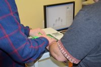 ręce dwóch mężczyzn położone na urządzeniu do pobierania odcisków palców. W tle widać monitor, na którym wyświetlają się odciski palców