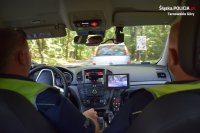 Zdjęcie kolorowe. Na fotografii widać wnętrze radiowozu i siedzących w nim dwóch policjantów. W tle przez przednią szybę radiowozu widać zatrzymany do kontroli samochód osobowy.