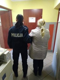 Pomieszczenie, w którym policjantka trzyma pod rękę zatrzymaną kobietę. Obie Panie są odwrócone tyłem
