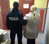 Pomieszczenie, w którym policjantka trzyma pod rękę zatrzymaną kobietę. Obie Panie są odwrócone tyłem