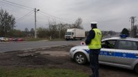 Na fotografii widać policjanta stojącego obok radiowozu. Mundurowy w dłoni trzyma tarczę służącą do zatrzymywania pojazdów. W tle fotografii widać jadący ulicą samochód ciężarowy.