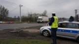 Na fotografii widać policjanta stojącego obok radiowozu. Mundurowy w dłoni trzyma tarczę służącą do zatrzymywania pojazdów. W tle fotografii widać jadący ulicą samochód ciężarowy.