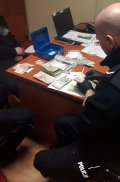 Na fotografii widać siedzącego przy biurku policjanta. Na biurku znajdują się pliki banknotów