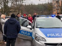 Na fotografii widać policyjny radiowóz,  obok którego stoją osoby niepełnosprawne i  ich opiekunowie. Wśród nich znajduje się dzielnicowy. W samochodzie otwarte są drzwi od strony pasażera