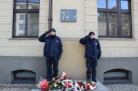 Przy murze Komendy, obok tablicy pamiątkowej stoją policjanci. Mundurowi oddają honor. Pd tablicą leżą wiązanki kwiatów oraz stoją zapalone znicze