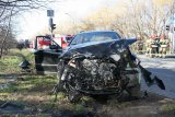 fotografia kolorowa: na zdjęciu widać rozbity przód samochodu osobowego - wyraźnie widać silnik pojazdu. W tle zdjęcia widać wozy strażackie i stojących na ulicy strażaków