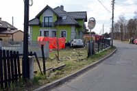 Na zdjęciu widać drogę i zniszczone ogrodzenie. W oddali widać dom jednorodzinny i &quot;wbity&quot; w ścianę budynku samochód osobowy. Obok samochodu widać czerwony parawan.