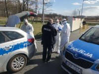 na fotografii widać policyjne radiowozy, obok których policjanci zakładają kombinezony ochronne