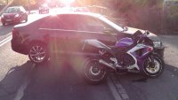 fotografia kolorowa: motocykl oparty o bok samochodu osobowego. W tle zdjęcia widać drogę, po której jadą samochody