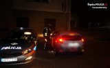 Na fotografii widać zaparkowany policyjny radiowóz oraz policjanta kontrolującego kierowcę samochodu osobowego