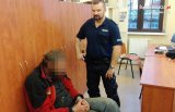 fotografia kolorowa: policjant stoi obok siedzącego na krześle, zatrzymanego mężczyzny.