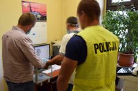fotografia kolorowa: pomieszczenie, w którym znajduje się dwóch policjantów oraz zatrzymany mężczyzna. Jeden z policjantów pobiera od zatrzymanego odciski palców, które widać na monitorze komputera.