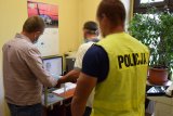 fotografia kolorowa: pomieszczenie, w którym znajduje się dwóch policjantów oraz zatrzymany mężczyzna. Jeden z policjantów pobiera od zatrzymanego odciski palców, które widać na monitorze komputera.