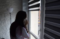 Przy oknie stoi młoda dziewczyna, która przy uchu trzyma telefon komórkowy.