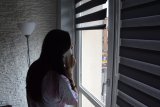 Przy oknie stoi młoda dziewczyna, która przy uchu trzyma telefon komórkowy.