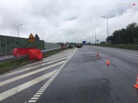 Na fotografii widać prosty odcinek autostrady z zabezpieczonym miejscem wypadku drogowego ze skutkiem śmiertelnym.  Z lewej strony zdjęcia widać parawan koloru czerwonego, a w oddali pojazdy służb ratowniczych.