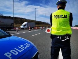 Na zdjęciu widać drogę w poprzek, której stoi policyjny radiowóz, a przy nim policjant, który trzyma w dłoni &quot;lizaka&quot; do zatrzymywania samochodów