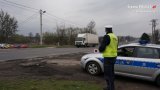 Na fotografii widać policjanta stojącego obok radiowozu. Mundurowy w dłoni trzyma tarczę służącą do zatrzymywania pojazdów. W tle fotografii widać jadący ulicą samochód ciężarowy.