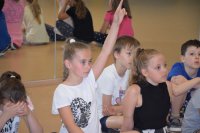 fotografia kolorowa: na podłodze w sali z lustrami, siedzi grupa dzieci. Jedna z dziewczynek ma podniesioną do góry rękę ponieważ chce zadać policjantom pytanie
