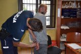 Na fotografii widać pomieszczenie, w którym policjant zapina kajdanki zatrzymanemu mężczyźnie