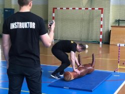Na fotografii widać salę gimnastyczną, na której policjant - instruktor, mierzy czas innemu mężczyźnie, który przenosi manekina