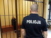 Na fotografii widać policjanta, który obserwuje mężczyznę siedzącego w policyjnej celi.