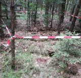 Zdjęcie przedstawia teren leśny, na którym leży pocisk przeciwpancerny. Teren wokół pocisku jest ogrodzony taśmą.