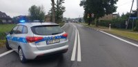 Na fotografii widać policyjny radiowóz zaparkowany na drodze.Pojazd ma włączone sygnały świetlne. W tle fotografii widać dużą grupę osób z transparentami.