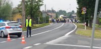 Na fotografii widać zaparkowany w poprzek drogi policyjny radiowóz. Obok widać policjanta w kamizelce.  W tle fotografii widać dużą grupę osób z transparentami.