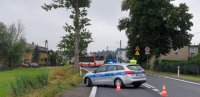 Na fotografii widać zaparkowany w poprzek drogi policyjny radiowóz. Obok widać policjanta w kamizelce.  W tle fotografii widać autobus i dużą grupę osób z transparentami.