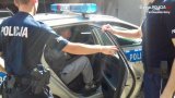 Na fotografii widać, jak dwóch policjantów otwiera drzwi radiowozu,  wewnątrz którego siedzi zatrzymany mężczyzna.