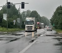 Na fotografii widać skrzyżowanie, na którym znajdują się samochody ciężarowe i osobowe. Przy jednej z ciężarówek stoi policjant i rozmawia z kierowcą