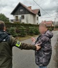 Na fotografii widać policjanta, który trzyma za rękę zatrzymanego mężczyznę. W oddali widać dom jednorodzinny.