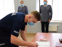 zdjęcie przedstawia policjanta składającego podpis pod ślubowaniem, w tle fotografii widać Naczelnika Wydziału Prewencji kom. Piotra Skolika oraz Specjalistę Zespołu Kadr i Szkolenia asp. szt. Anetę Bartoszewską.