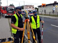 zdjęcie przedstawia dwóch policjantów stojących obok urządzenia służącego do wykonywania pomiarów. W tle widać wozy strażackie oraz karetki pogotowia.