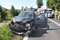 Na zdjęciu widać zniszczony samochód osobowy, zaparkowany na poboczu drogi. W tle zdjęcia widać karetki pogotowia, strażaków oraz policjanta