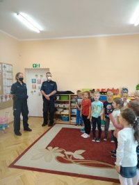 Na zdjęciu widać szkolną klasę, w której przed grupą uczniów stoi policjant i policjantka