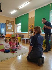 Na zdjęciu widać szkolną klasę, w której na podłodze siedzi grupka dzieci. Przed nimi klęczy policjantka, a obok stoi policjant.