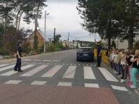 Na zdjęciu widać przejście dla pieszych, na środku którego stoi policjantka. Przed przejściem stoi grupa dzieci