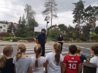 Na zdjęciu widać grupę dzieci odwróconą tyłem. Dzieci stoją przed przejściem dla pieszych, na środku którego stoi policjant i policjantka.