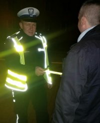 Na zdjęciu widać policjanta z Wydziału Ruchu Drogowego, ubranego w kamizelkę odblaskową, która świeci kiedy padają na nią światła samochodu. Policjant wręcza odblaskową opaskę pieszemu