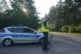 Na zdjęciu widać drogę biegnącą przez teren leśny. Przy drodze widać policjanta z Wydziału Ruchu Drogowego, który w dłoni trzyma urządzenie do badania stanu trzeźwości.