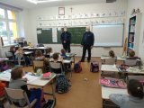 Na zdjęciu widać szkolną klasę, w której w ławkach siedzą uczniowie, a pod tablicą stoją dwaj policjanci