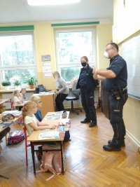 Na zdjęciu widać szkolną klasę, w której w ławkach siedzą uczniowie, a pod tablicą stoją policjantka i policjant.