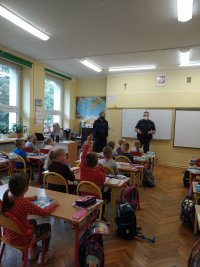 Na zdjęciu widać szkolną klasę, w której w ławkach siedzą uczniowie, a pod tablicą stoją policjantka i policjant. Jedno z dzieci podniosło rękę w górę chcąc zadać pytanie.