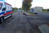 Zdjęcie przedstawia miejsce wypadku drogowego. Na ulicy leży rozbity motocykl, w poprzek drogi stoi policyjny radiowóz, a przed nim policjantka. Z lewej strony widać karetkę pogotowia, a z prawej, na parkingu drugi policyjny radiowóz i samochody osobowe.