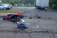 Zdjęcie przedstawia miejsce wypadku drogowego. Na ulicy leży rozbity motocykl. Na parkingu znajdującym się obok ulicy widać samochody osobowe oraz samochód ciężarowy, który brał udział w wypadku.