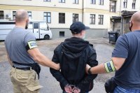 Na zdjęciu widać dwóch policjantów prowadzących zatrzymanego mężczyznę. Mężczyzna ma zapięte kajdanki na rękach trzymanych z tyłu.