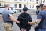 Na zdjęciu widać dwóch policjantów prowadzących zatrzymanego mężczyznę. Mężczyzna ma zapięte kajdanki na rękach trzymanych z tyłu.